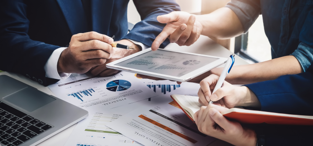 Three people sitting at a conference table with pens, legal pads, and tablets collaborating on financial data statements.